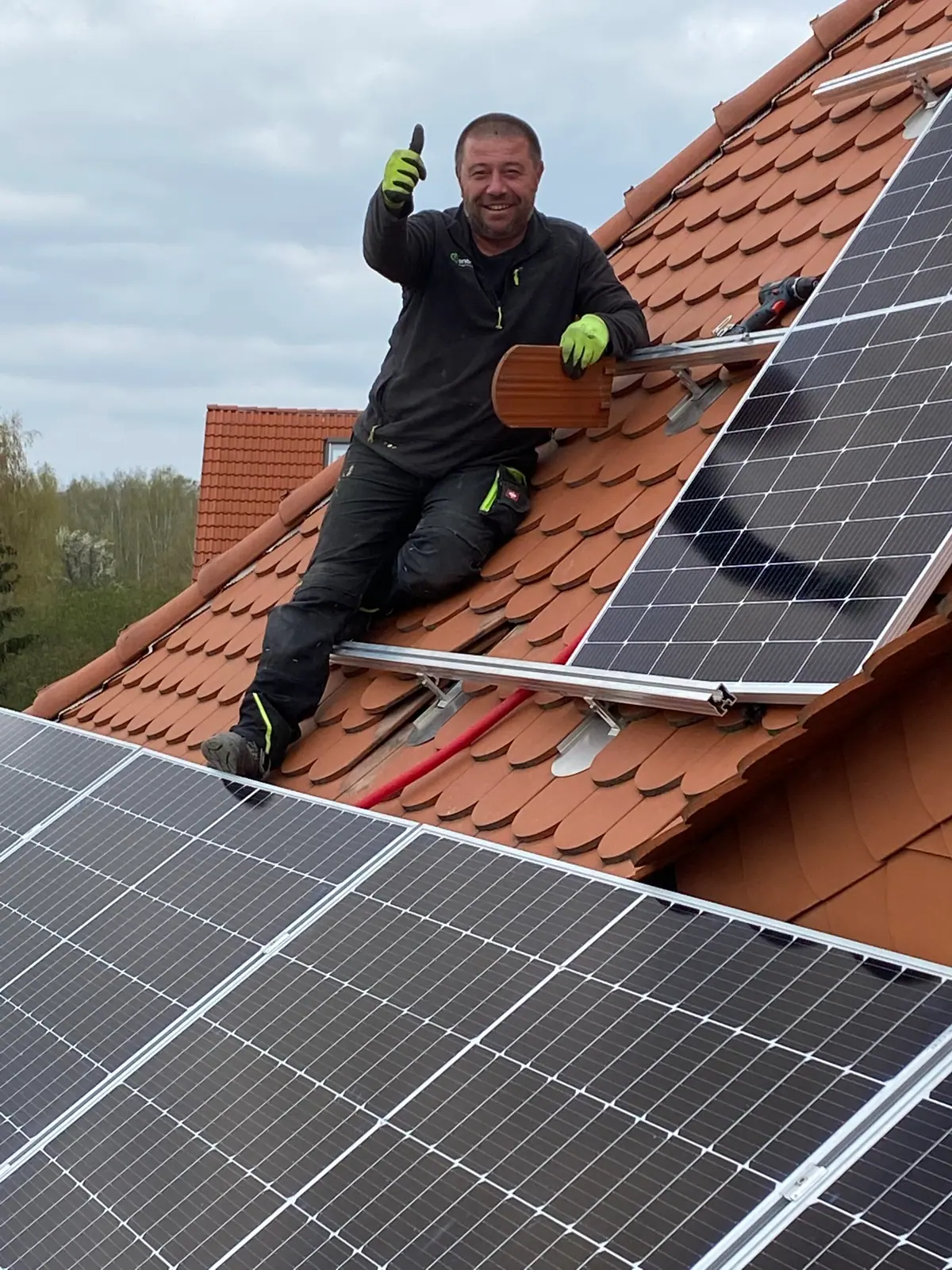 Monteur von enerbloc kniet zwischen Solarmodulen auf einem Ziegeldach und zeigt den Daumen nach oben